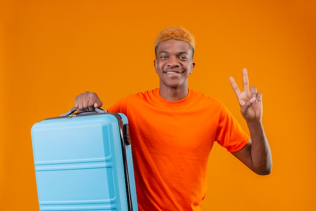 Ragazzo giovane viaggiatore che indossa la maglietta arancione che tiene la valigia sorridente che mostra il numero due o il segno di vittoria in piedi sopra la parete arancione