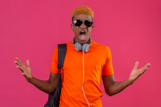 Young traveler boy wearing orange t-shirt with headphones and backpack crazy and mad shouting with angry expression on face with raised hands standing over pink wall