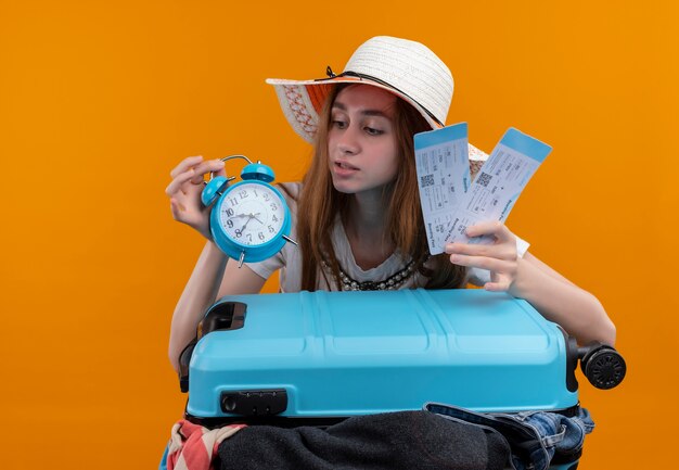 Young traveler girl holding airplane tickets and alarm clock with suitcase and looking at alarm clock on isolated orange wall
