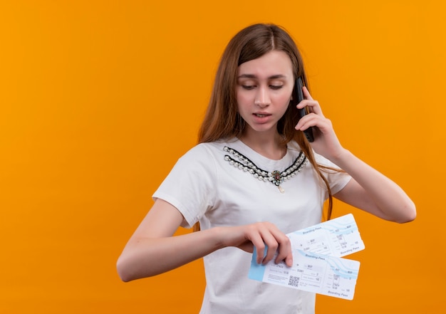 Бесплатное фото Молодая девушка путешественника разговаривает по телефону и держит билеты на самолет, глядя на изолированную оранжевую стену с копией пространства