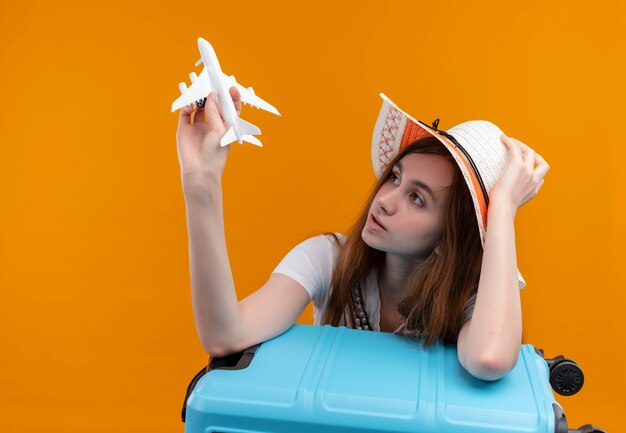 模型飛行機を保持し、それを見て、スーツケースに腕を置き、コピースペースのある孤立したオレンジ色の壁に帽子をかぶった若い旅行者の女の子