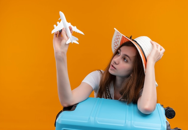 Young traveler girl wearing hat holding model plane and looking at it and putting arm on suitcase and hand on hat on isolated orange wall with copy space