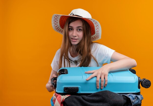 Young traveler girl wearing hat holding suitcase full of cloths looking  on isolated orange wall with copy space