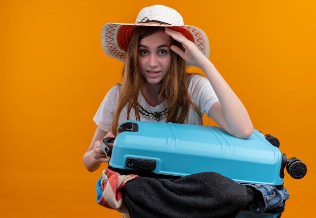 Young traveler girl wearing hat holding suitcase full of cloths and putting hand on head looking  on isolated orange wall with copy space