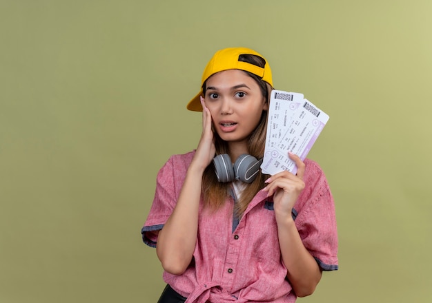 Free photo young traveler girl wearing pink shirt in cap with headphones around neck holding air tickets looking amazed and surprised with hand on face