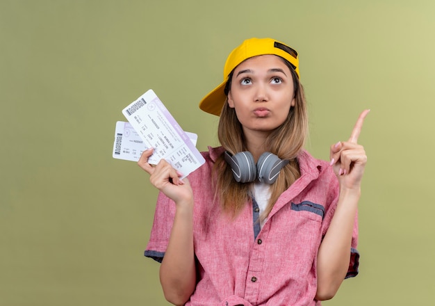 Young traveler girl wearing pink shirt in cap with headphones around neck holding air tickets looking up puzzled pointing finger up