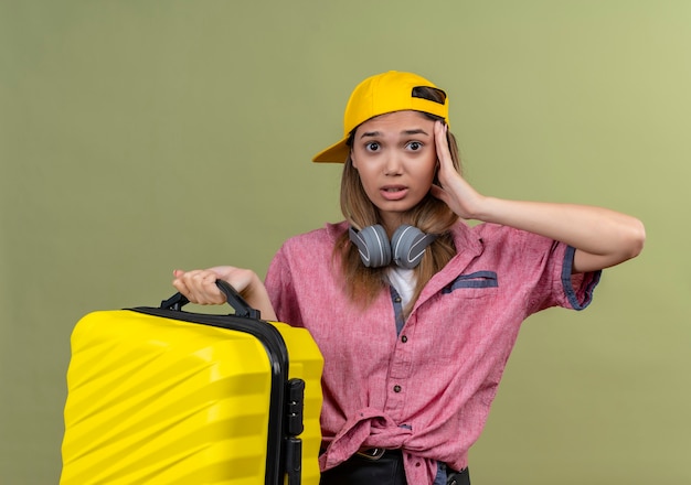 머리에 손으로 혼란과 매우 불안 찾고 가방을 들고 목에 헤드폰으로 모자에 분홍색 셔츠를 입고 젊은 여행자 소녀
