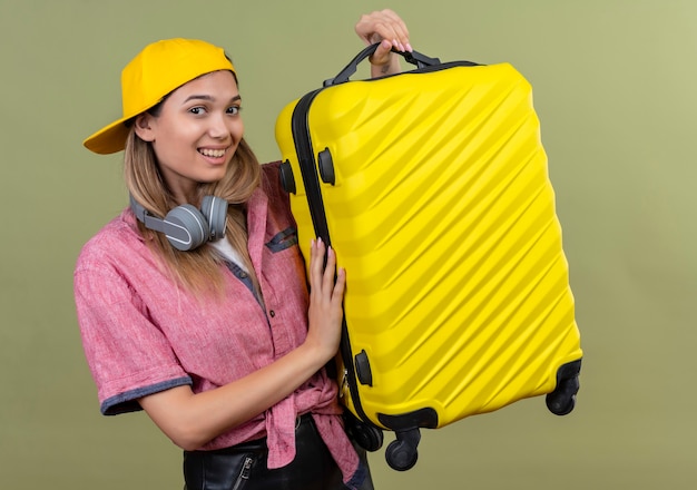 帽子をかぶったピンクのシャツを着て、首にヘッドフォンを持ってスーツケースを持って前向きで幸せな旅行の準備ができて笑顔