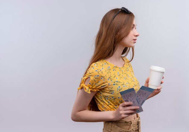 Young traveler girl wearing sunglasses on head holding airplane tickets looking at right side on isolated white wall with copy space