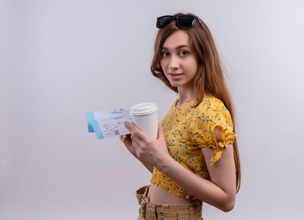 Young traveler girl wearing sunglasses on head holding airplane tickets and plastic coffee cup on isolated white wall with copy space