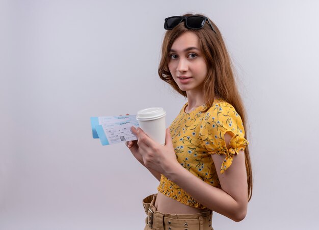 コピースペースと隔離された白い壁に飛行機のチケットとプラスチック製のコーヒーカップを保持している頭にサングラスをかけている若い旅行者の女の子