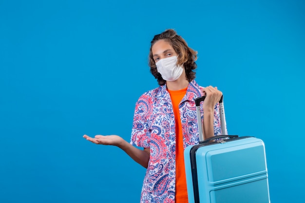 Young traveler guy wearing facial protective mask holding travel suitcase clueless and confused standing with arm raised