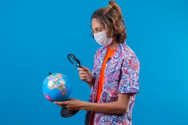 Young traveler guy wearing facial protective mask holding travel suitcase holding and looking through magnifying glass on globe with interest standing over blue background