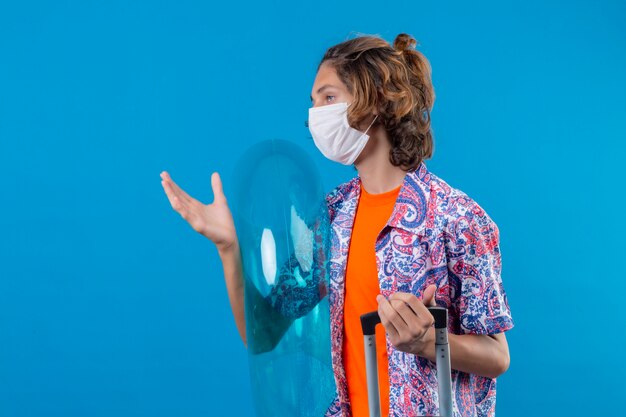 Young traveler guy wearing facial protective mask holding travel suitcase and inflatable ring looking aside with serious confident expression standing with arm raised over blue background
