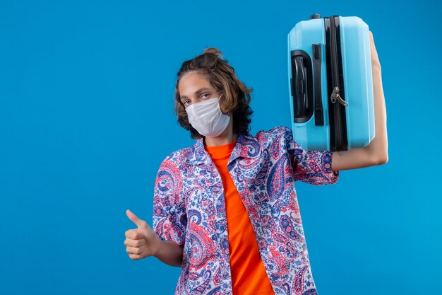 Young traveler guy wearing facial protective mask holding travel suitcase looking confident showing thumbs up standing over blue background