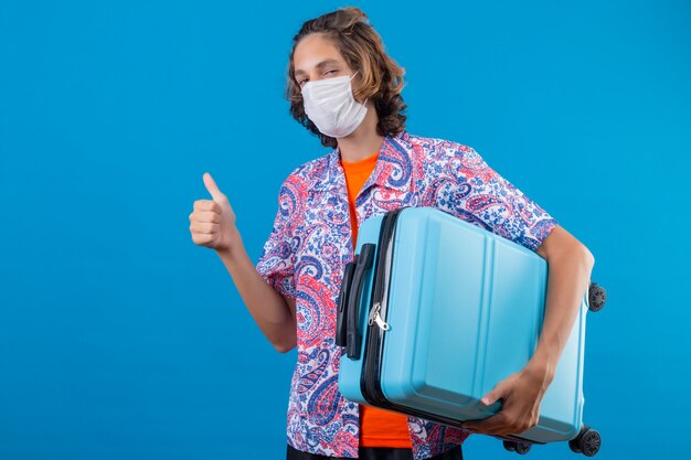 Young traveler guy wearing facial protective mask holding travel suitcase looking confident showing thumbs up standing