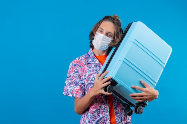 Young traveler guy wearing facial protective mask holding travel suitcase looking positive and happy standing over blue background