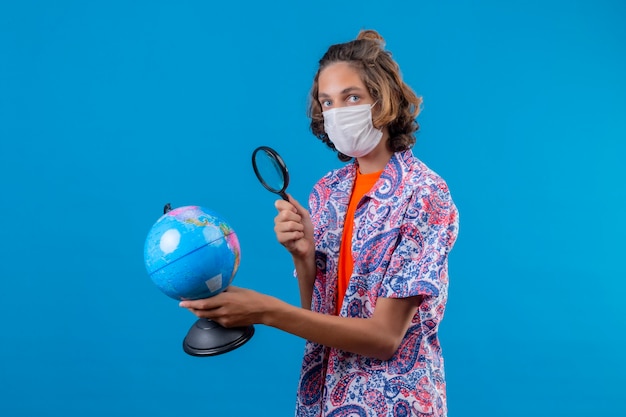 Ragazzo giovane viaggiatore che indossa la maschera protettiva per il viso tenendo la valigia da viaggio e guardando attraverso la lente di ingrandimento sul globo con interesse in piedi su sfondo blu