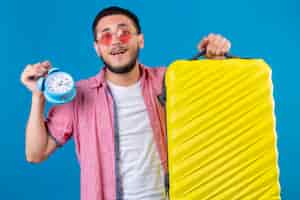 Бесплатное фото Молодой путешественник красивый парень в солнцезащитных очках, держит дорожный чемодан и будильник, выглядит счастливым и позитивным на синем фоне