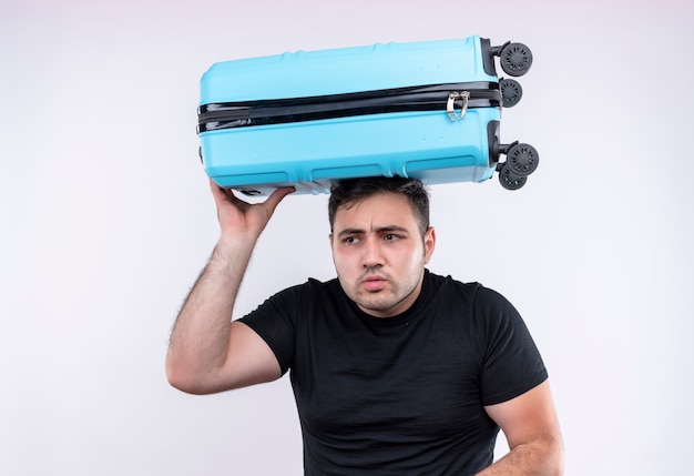 Uomo giovane viaggiatore in maglietta nera che tiene la valigia sulla sua testa che osserva da parte in piedi preoccupato sopra il muro bianco