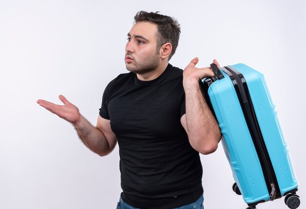 Young traveler man in black t-shirt holding suitcase looking aside confused and very anxious standing over white wall