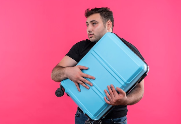 Young traveler man in black t-shirt holding suitcase looking aside confused with fear expression standing over pink wall