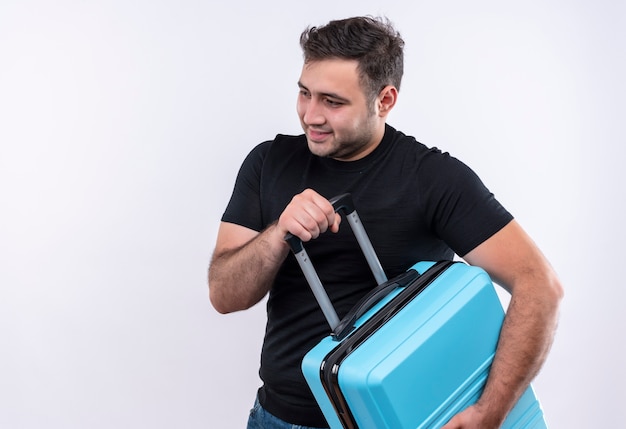 Young traveler man in black t-shirt holding suitcase looking aside positive and happy smiling standing over white wall