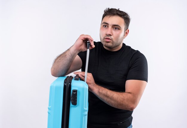 Young traveler man in black t-shirt holding suitcase looking aside puzzled standing over white wall