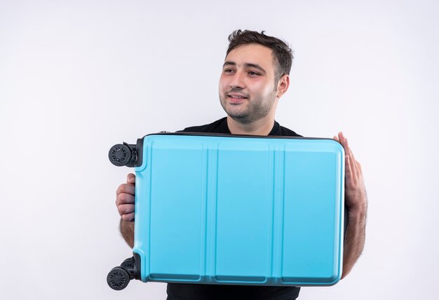 Young traveler man in black t-shirt holding suitcase looking aside smiling with happy face standing over white wall