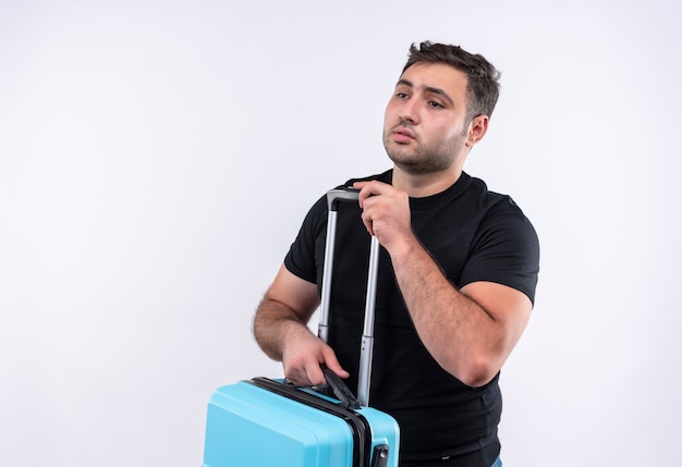 Young traveler man in black t-shirt holding suitcase looking aside with sad expression on face standing over white wall