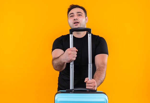 Young traveler man in black t-shirt holding suitcase looking confident, ready for vacation standing over orange wall