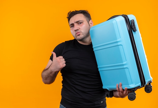 Young traveler man in black t-shirt holding suitcase looking confused pointing back with thumb standing over orange wall