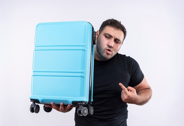 Young traveler man in black t-shirt holding suitcase pointing with index finger to it looking aside anxious standing over white wall