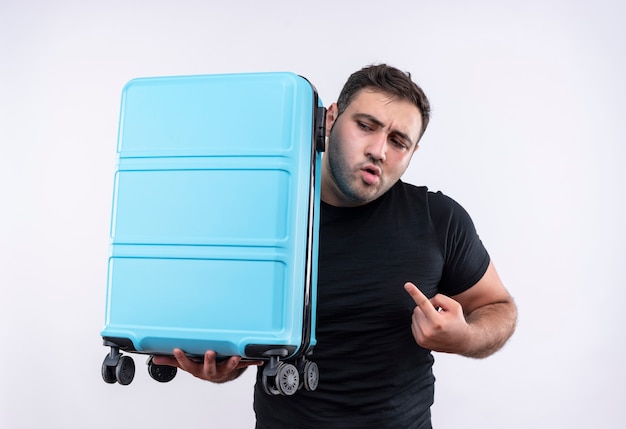 人差し指でスーツケースを持って白い壁の上に立っている不安を脇に見ている黒いTシャツを着た若い旅行者の男