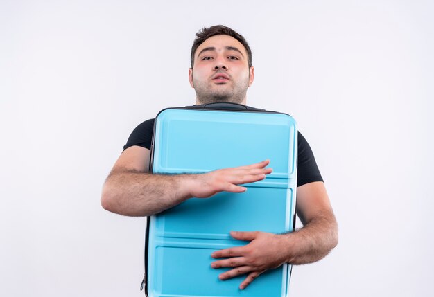 Young traveler man in black t-shirt holding suitcase positive and happy standing over white wall