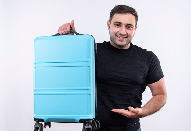 Young traveler man in black t-shirt holding suitcase presenting it with arm smiling confident standing over white wall