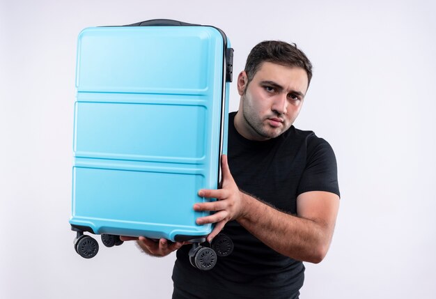 白い壁の上に立って困惑したスーツケースを保持している黒いTシャツの若い旅行者の男