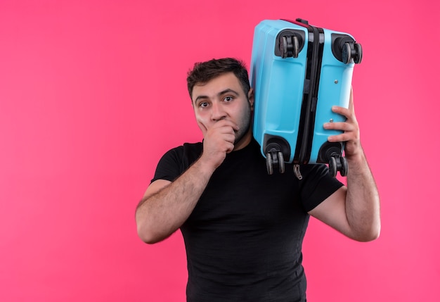 Young traveler man in black t-shirt holding suitcase shocked standing over pink wall