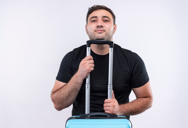 Young traveler man in black t-shirt holding suitcase with confident smile on face standing over white wall