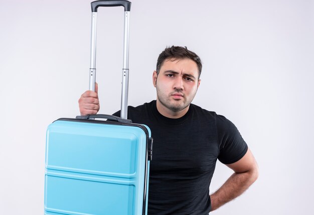 Young traveler man in black t-shirt holding suitcase with frowning face standing over white wall