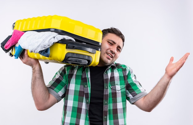 Young traveler man in checked shirt holding suitcase full of clothes looking disappointed and confused standing over white wall