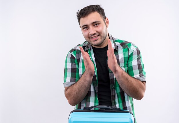 Young traveler man in checked shirt holding suitcase happy and positive smilin and applauding standing over white wall