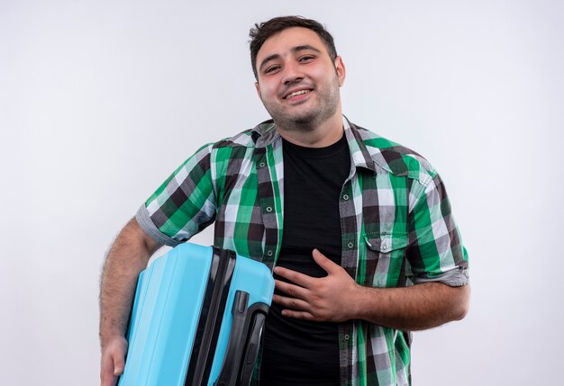 Young traveler man in checked shirt holding suitcase happy and positive smiling standing over white wall