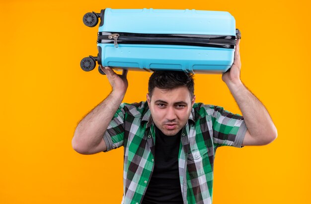 Young traveler man in checked shirt holding suitcase on his head looking tired with annoyed expression standing over orange wall