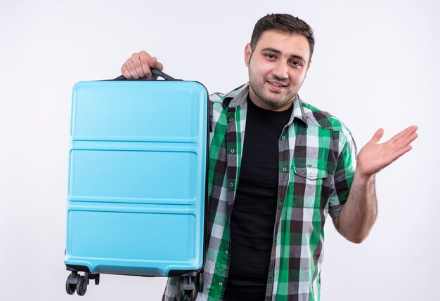Young traveler man in checked shirt holding suitcase looking confused spreading with arm to the side smiling standing over white wall
