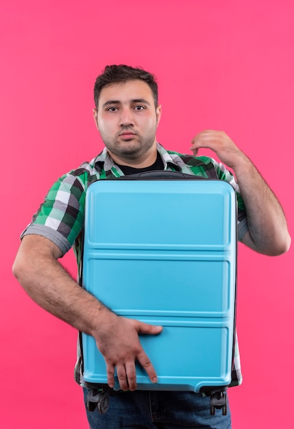 Uomo giovane viaggiatore in camicia a quadri che tiene la valigia cercando confuso e molto ansioso in piedi oltre il muro rosa