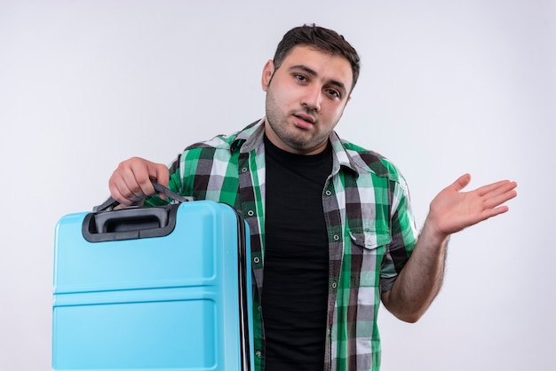 Young traveler man in checked shirt holding suitcase looking uncertain and confused spreading with arm to the side standing over white wall