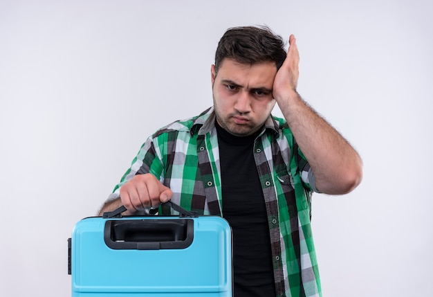 Young traveler man in checked shirt holding suitcase looking uncertain and confused touching his head blowing cheeks standing over white wall