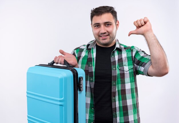 Young traveler man in checked shirt holding suitcase pointing with finger to himself with confident smile on face standing over white wall