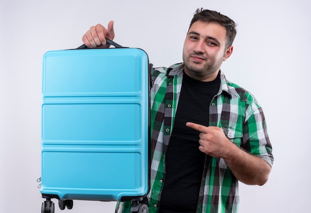 Young traveler man in checked shirt holding suitcase pointing with index finger to it with confident smile on face standing over white wall